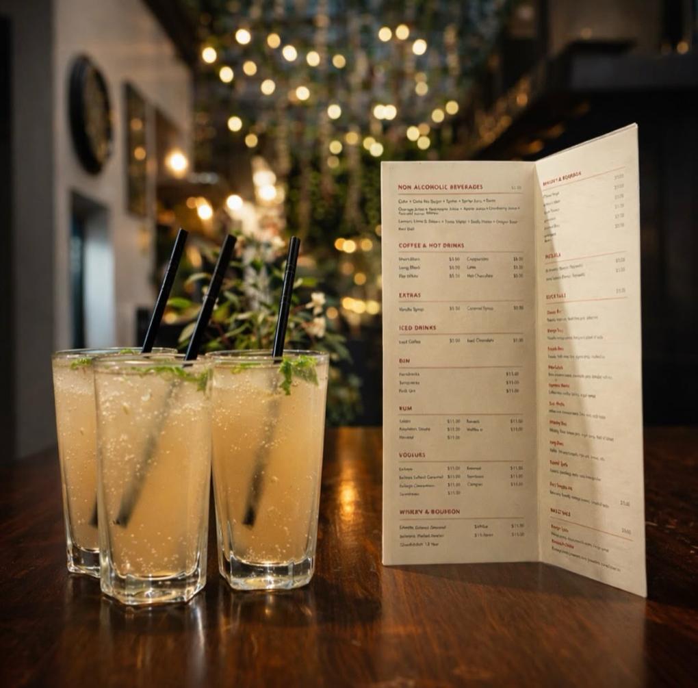Three tall drinks beside the open drinks list, with string lights and greenery blurred behind.