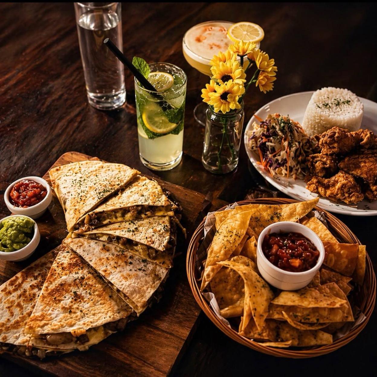 A moody table of quesadillas on a board, corn chips with salsa, fried chicken with rice, and cocktails.