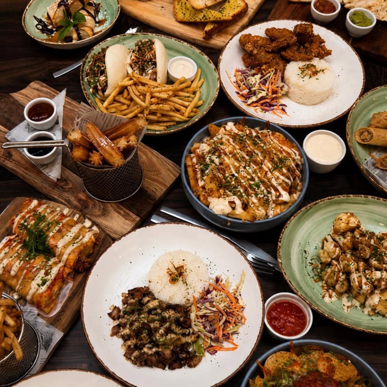 An overhead spread of bao, fried chicken, loaded fries, churros, sisig, lumpia and enchiladas across a dark table.