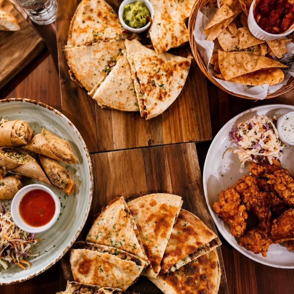Overhead view of quesadillas on boards, lumpia, fried chicken with slaw and a basket of corn chips.