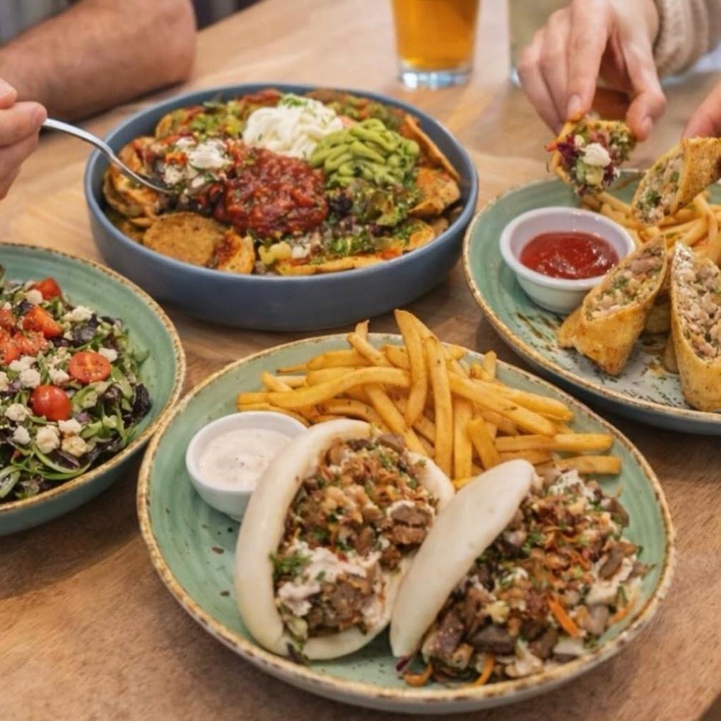 Hands reaching across a table of nachos, bao buns, salad and a wrap.